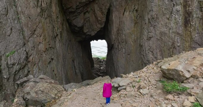 Pink jacket woman look at ocean through Torghatten mountain hole; drone reveal
