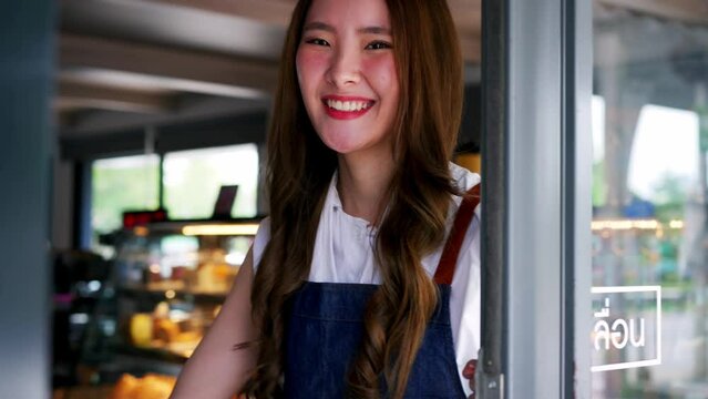 Young adult Asian female barista welcoming a customer walking into the coffee shop with a friendly smile and warm welcome. Small Business owner in Asia.