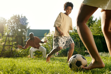 Legs of barefoot girl kicking soccer ball to her brother while playing on green lawn with him and their father standing on background - Powered by Adobe