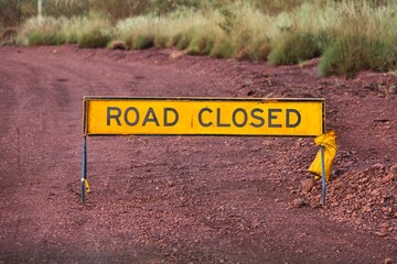 A yellow sign saying 'road closed'. Road closed sign on an unpaved road. Yellow construction site sign. Warning signage on a red-colored gravel road. Western Australia gravel highway closed.   