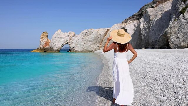 A elegant tourist woman in a white summer dress enjoys the beautiful beach of Lalaria, Skiathos island, Greece, during sunset time