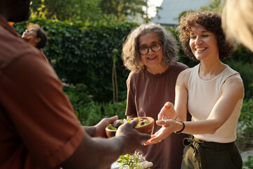 Focus on young cheerful brunette woman taking bowl with food from hands of her husband helping her...