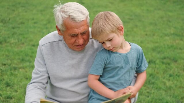 Happy grandfather reading book to curious grandson outdoors. Close up. Grandpa with grandchild spending time together. Family time comes in various forms. 4s year boy and senior man read story in park