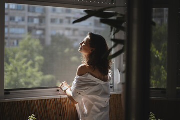 Young smiling caucasian woman in a white shirt standing on the balcony in the morning meeting a new day. View of summer city