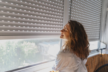 Young woman opening roller blind shutters on the balcony on a summer day - 629857538
