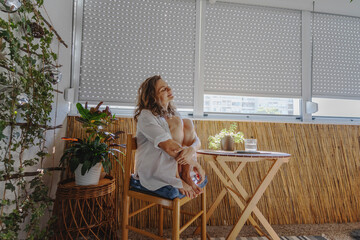 Young caucasian woman in a white shirt sitting on a balcony with roller shutters. Sun protection...