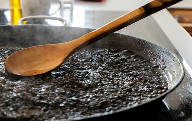 Black rice with squid being cooked in a paella pan and a wooden spoon. Black rice with cuttlefish. Rice al nero di sepia. Black risotto