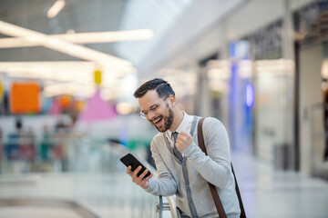 A young elegant man is standing at the shopping mall and celebrating his paycheck while holding his phone and laughing at it.