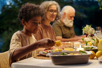 Cute little boy taking homemade food from container and putting it on his plate while sitting by served table during outdoor family dinner