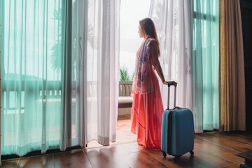 Traveler woman in pink dress with suitcase standing in room her hand opening curtains looking out...