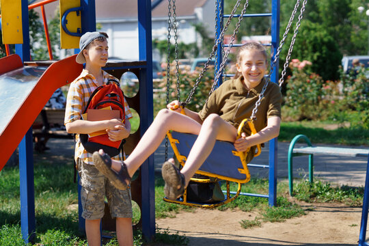 schoolchildren, a teenage boy and a girl play on the playground in the city park after school, a girl rides on a swing, a bright summer day