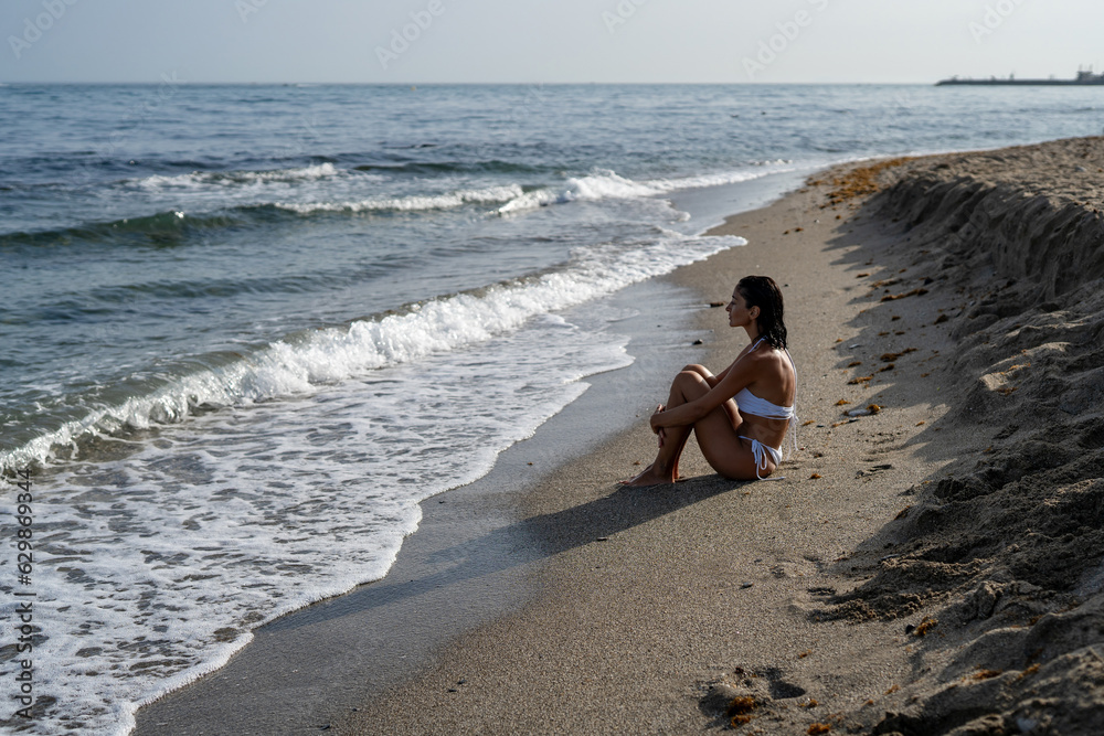 Wall mural beautiful brunette girl in a white swimsuit sits on the sand on the seashore