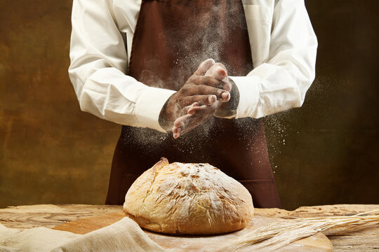 Unrecognizable baker in bround apron pouring flour on round loaf of bread over brown background.