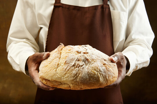 Unrecognizable baker in bround apron holding round loaf of bread in hands over brown background.