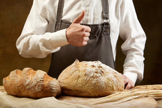 Unrecognizable baker in grey apron likes round bread and long loaf over brown background.