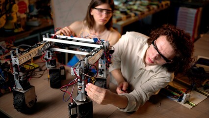 Two young engineers fixing a mechanical robot with wheels in the workshop, wearing industrial glasses