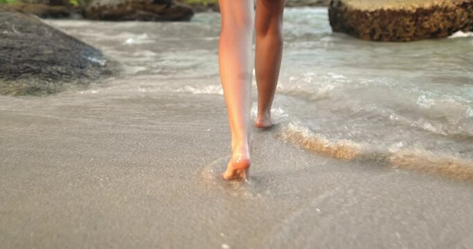 Woman legs bare foot walk sand beach, make sea water splashes. Slim female feet run at seaside surf in evening light. Girl tourist on summer vacation on tropical island resort. Slow motion, back view