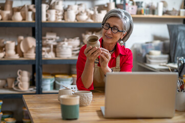 Mature woman painting pottery earthenware product in front of laptop while live streaming.