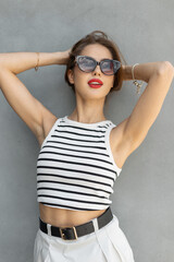 Beautiful young fashion girl with fashionable sunglasses in casual summer outfit stands near a gray wall on the street
