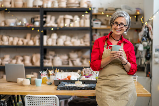 Happy mature female pottery master taking coffee break in ceramics studio.