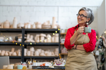 Senior craftswoman resting at pottery workshop drinking coffee.