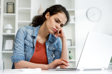 Young curly brunette, sitting stressed at the table and working on a laptop in a bright home office, apathy and depression from overwork at work. Burnout from the modern world. Programmer, student