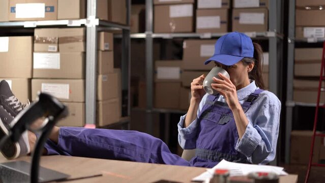 Warehouse worker woman has a break drinks coffee or tea while relaxing at the table