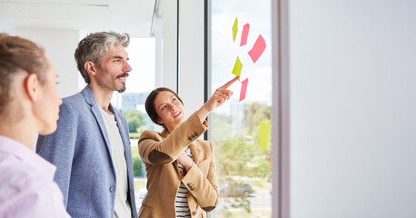 Happy businessman discussing over sticky notes on window with female colleagues during meeting in office