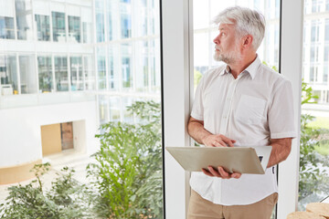 Thoughtful senior businessman with laptop looking out of window while leaning in office lounge