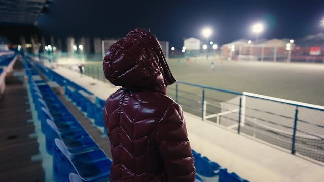 Side angle view Caucasian woman sitting at night on stadium tribune as blurred players playing football at background. Lady enjoying hobby outdoors
