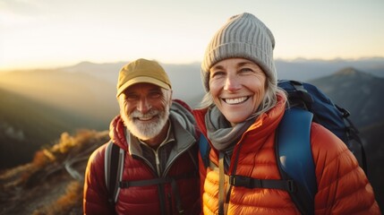 An active senior couple hiking in the mountains wearing beanies, puffer jackets, and backpacks at dawn, smiling. Mountains are in the background and the sun is rising in the horizon.