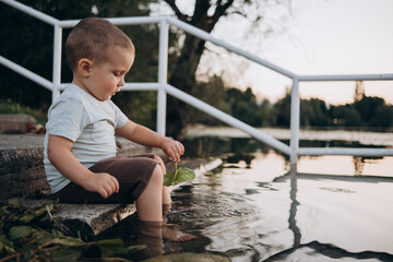  child sits on the steps on the shore of the lake and holds a leaf of a water lily in his hands