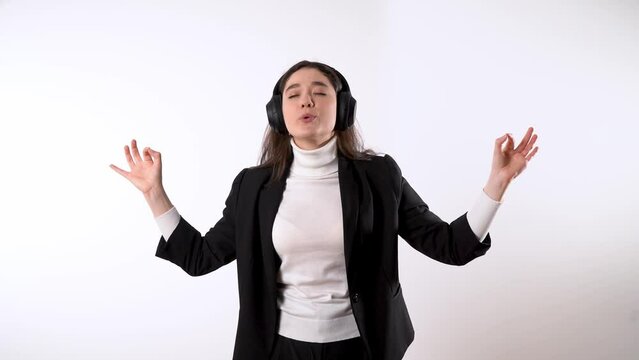 Tired looking smartly dressed young brunette against white background putting headphones with relaxing music and making OMM meditation sign with her fingers 