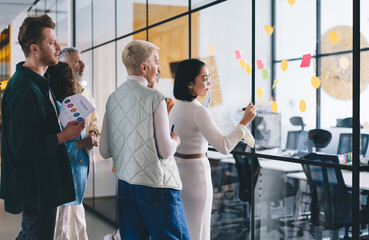 Group of experienced office workers talking while collaborating on informative memo sticked to glass board in office interior, confident male and female partners briefing and communicating in workshop