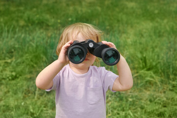 Little girl using binoculars in a forest. 2 years kid looking ahead. Smiling baby with spyglass. Adventure, Imagination, travel concept. Freedom, vacation. Happy child playing outdoor in summer field