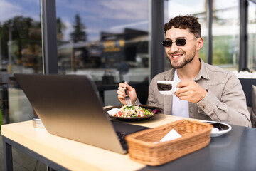 Handsome young man working on laptop and smiling while sitting in sidewalk cafe