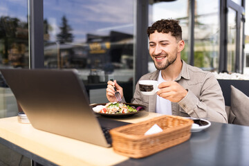 Handsome young man working on laptop and smiling while sitting in sidewalk cafe