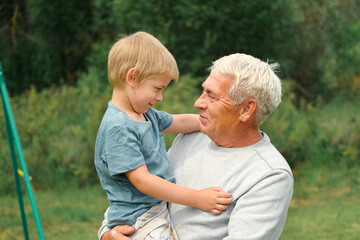 Fototapeta premium Grandfather and grandchild baby have fun during walk In Park. Happy family time. Old man grandpa hugging 4 years child boy at summer day. Smiling Senior male spending time with his grandson together.