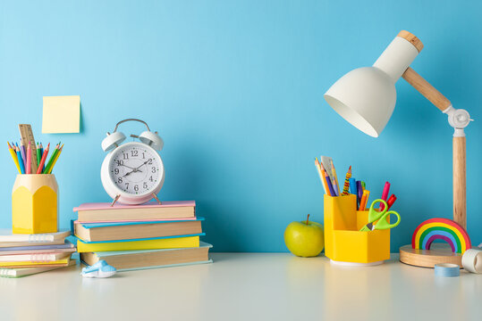 Classroom magic on display: side view photo of desk adorned with school essentials, pencil holders, pens, ruler, books, copybooks, alarm clock, lamp, apple, sticky notes on a blue wall backdrop