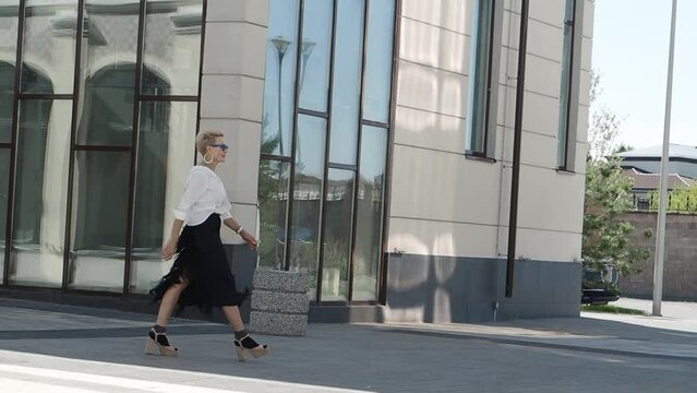 Confidence and Beautiful blonde woman with stylish short haircut walks city streets in fashionable chic white shirt and black fringed skirt with high heeled sandals and socks, fashion icon of style.
