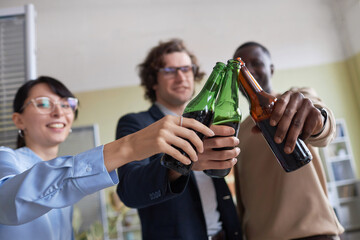 Closeup of colleagues clinking beer bottles while celebrating in office at workplace party, copy space