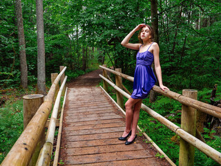 Young teenager girl dressed in stylish blue high fashion dress posing on a wooden bridge in a forest park. Prom time. Model wears design clothes. Glamour and elegance concept.