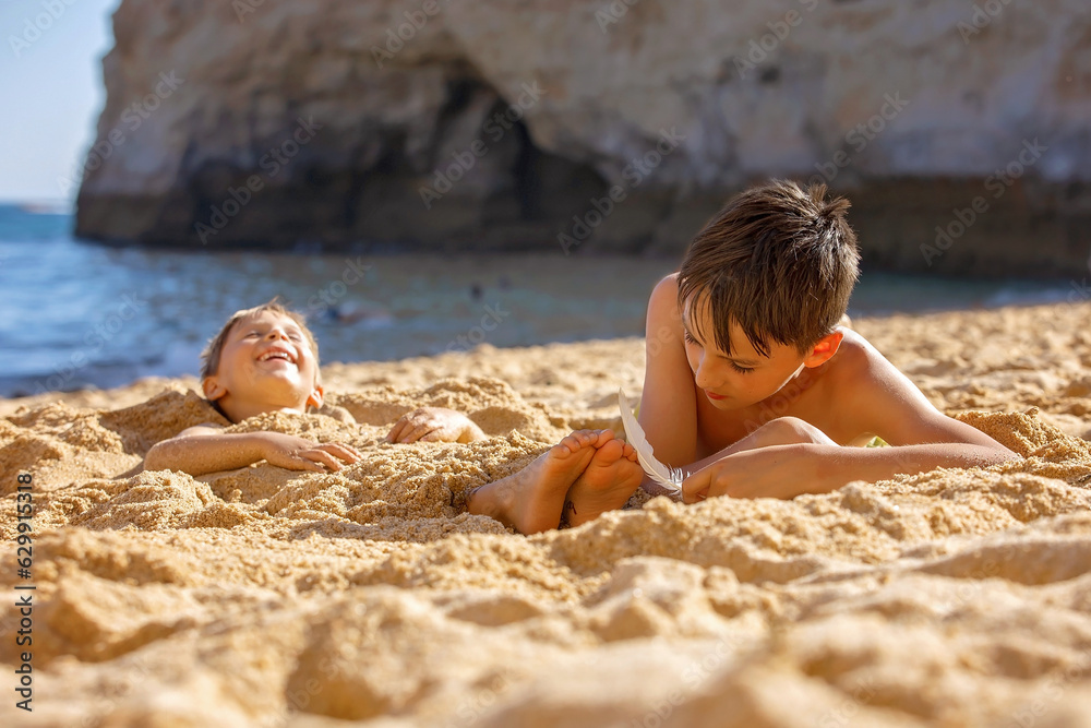 Wall mural child, tickling sibling on the beach on the feet with feather, kid cover in sand, smiling, laughing