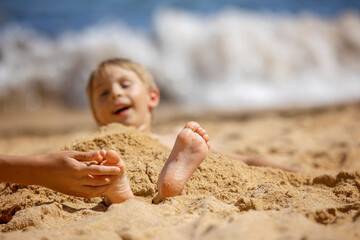 Child, tickling sibling on the beach on the feet with feather, kid cover in sand, smiling, laughing