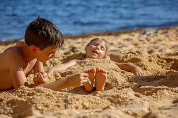 Child, tickling sibling on the beach on the feet with feather, kid cover in sand, smiling, laughing