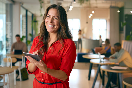 Mature Businesswoman Standing In Busy Office Using Digital Tablet