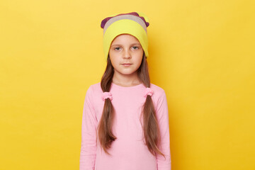 Serious confident little girl with ponytails wearing pink shirt and beanie hat posing isolated over...
