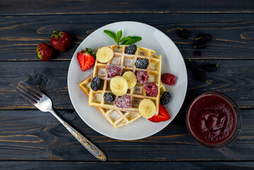 Belgian waffles with summer berries and powdered sugar in a white plate on a dark wooden background.