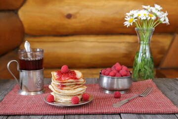 On a wooden table there is a stack of pancakes with fresh raspberries and jam near a bowl of ripe berries, a glass of tea and a vase of flowers.