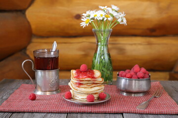 Obraz na płótnie Canvas On a wooden table there is a stack of pancakes with fresh raspberries and jam near a bowl of ripe berries, a glass of tea and a vase of flowers.